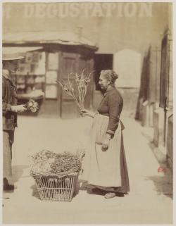 une femme vêtue d'un tablier balnc vend des fleurs à un jeune homme